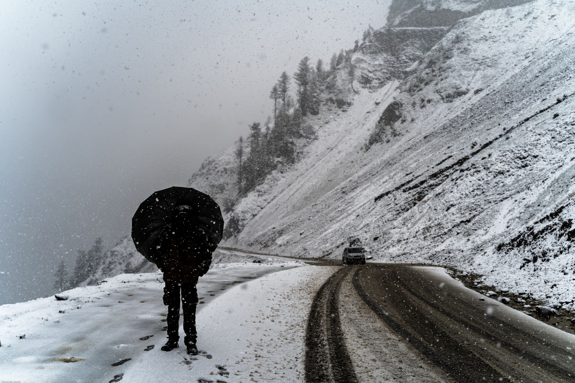A person with an umbrella walks a snowy mountain road as snow falls heavily.