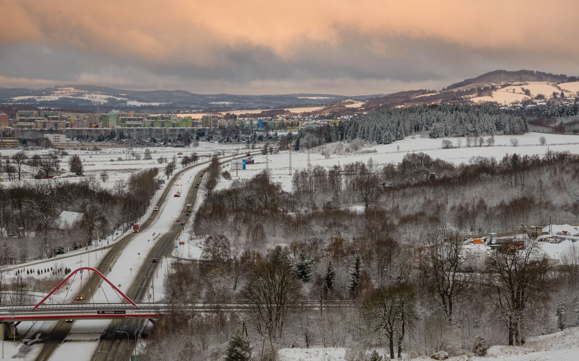 Aerial view of a snowy landscape featuring a bridge, roads, and a distant town under a pastel sky.