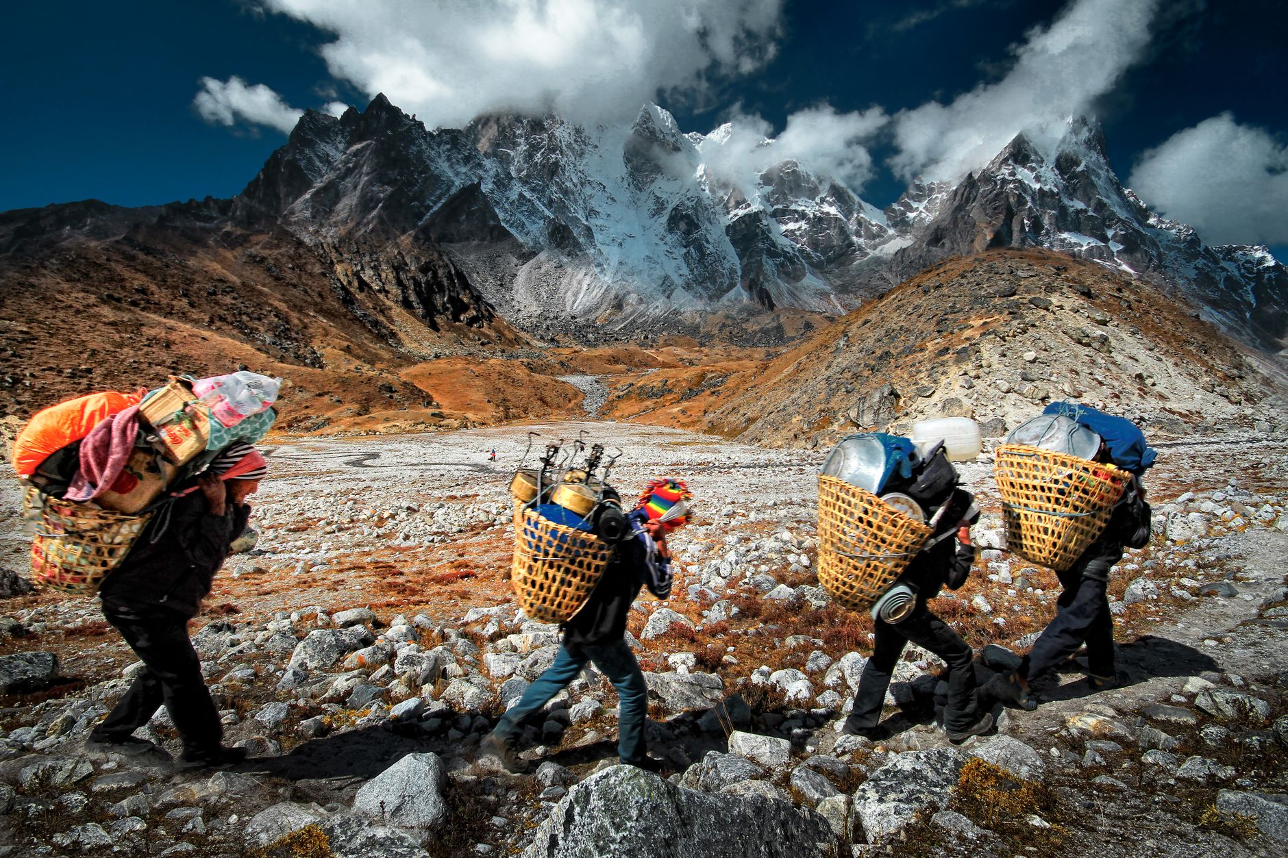 Porters carrying goods through rugged Himalayan terrain in Nepal with dramatic mountain backdrop.