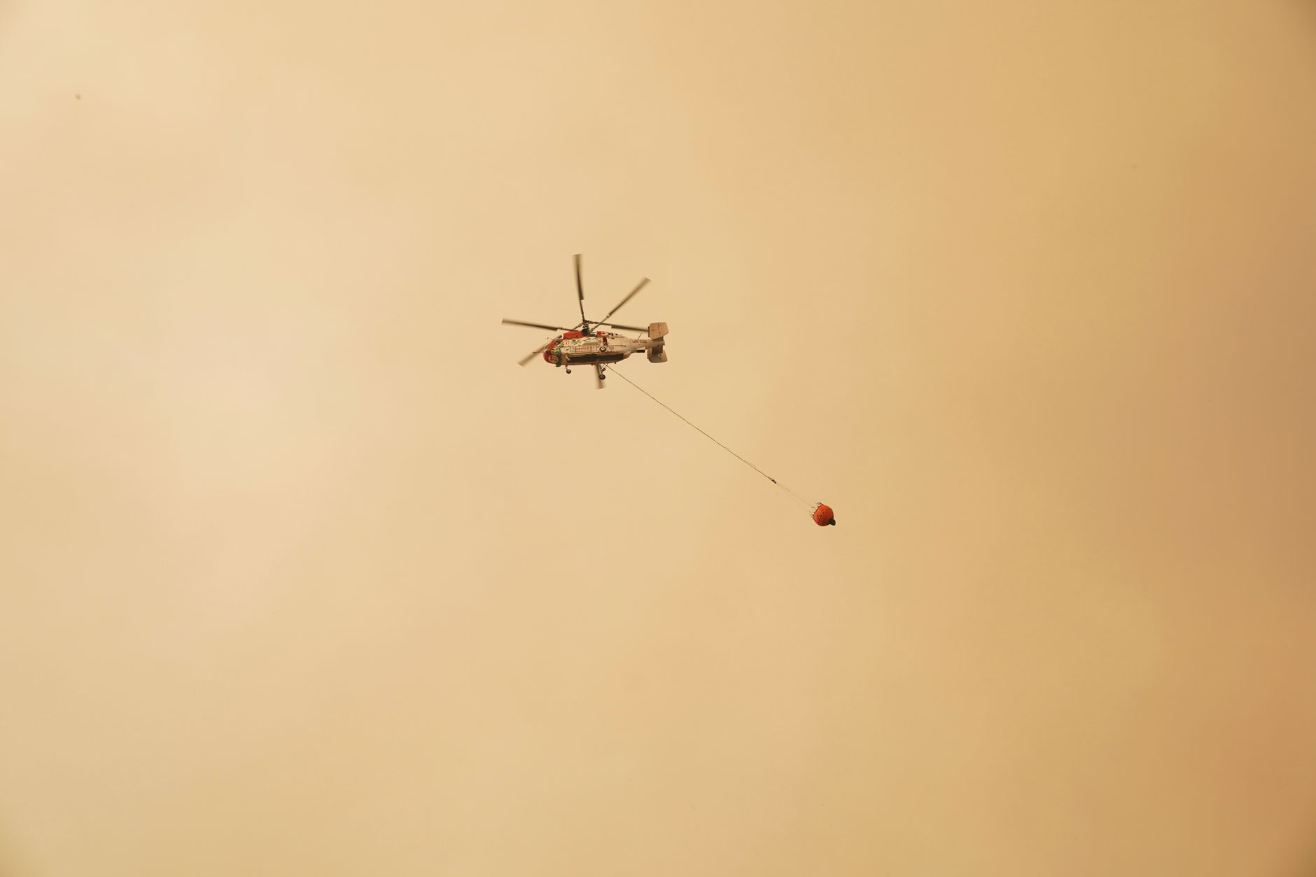 Helicopter carries water bucket against hazy sky, illustrating aerial firefighting during a wildfire.