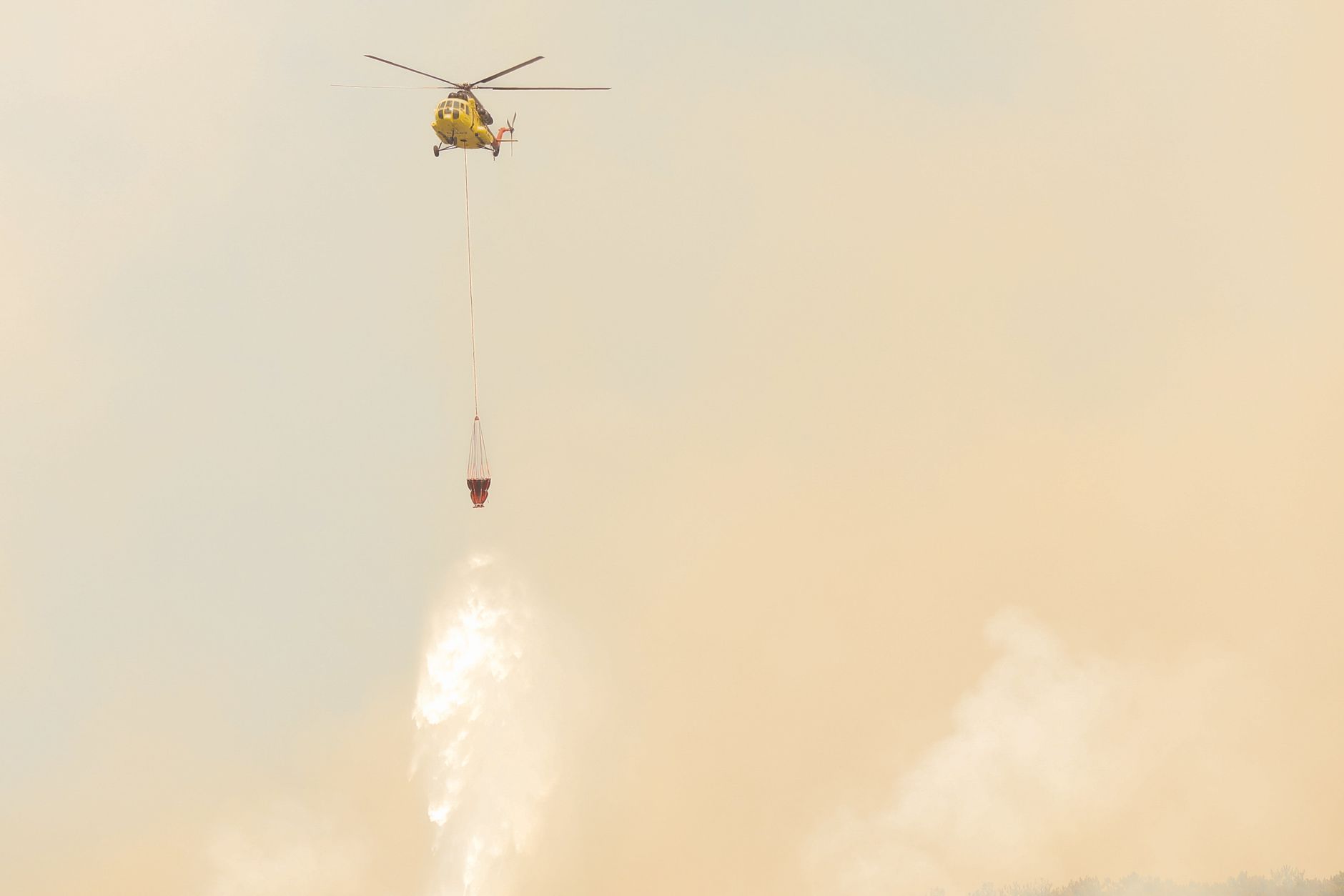 Yellow helicopter drops water to combat wildfires amidst smoky skies.