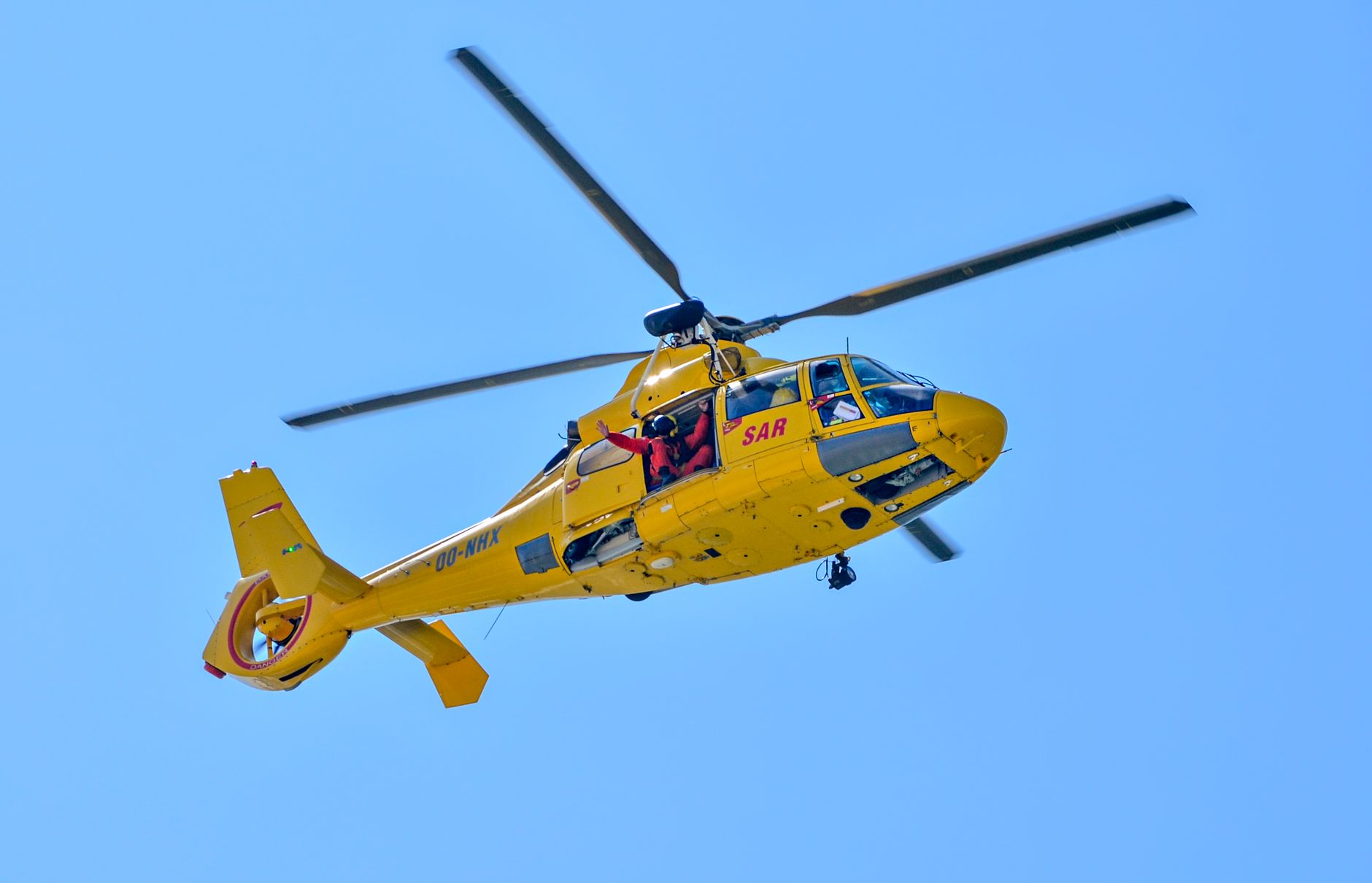 A bright yellow SAR helicopter mid-air, performing a mission under clear blue skies.