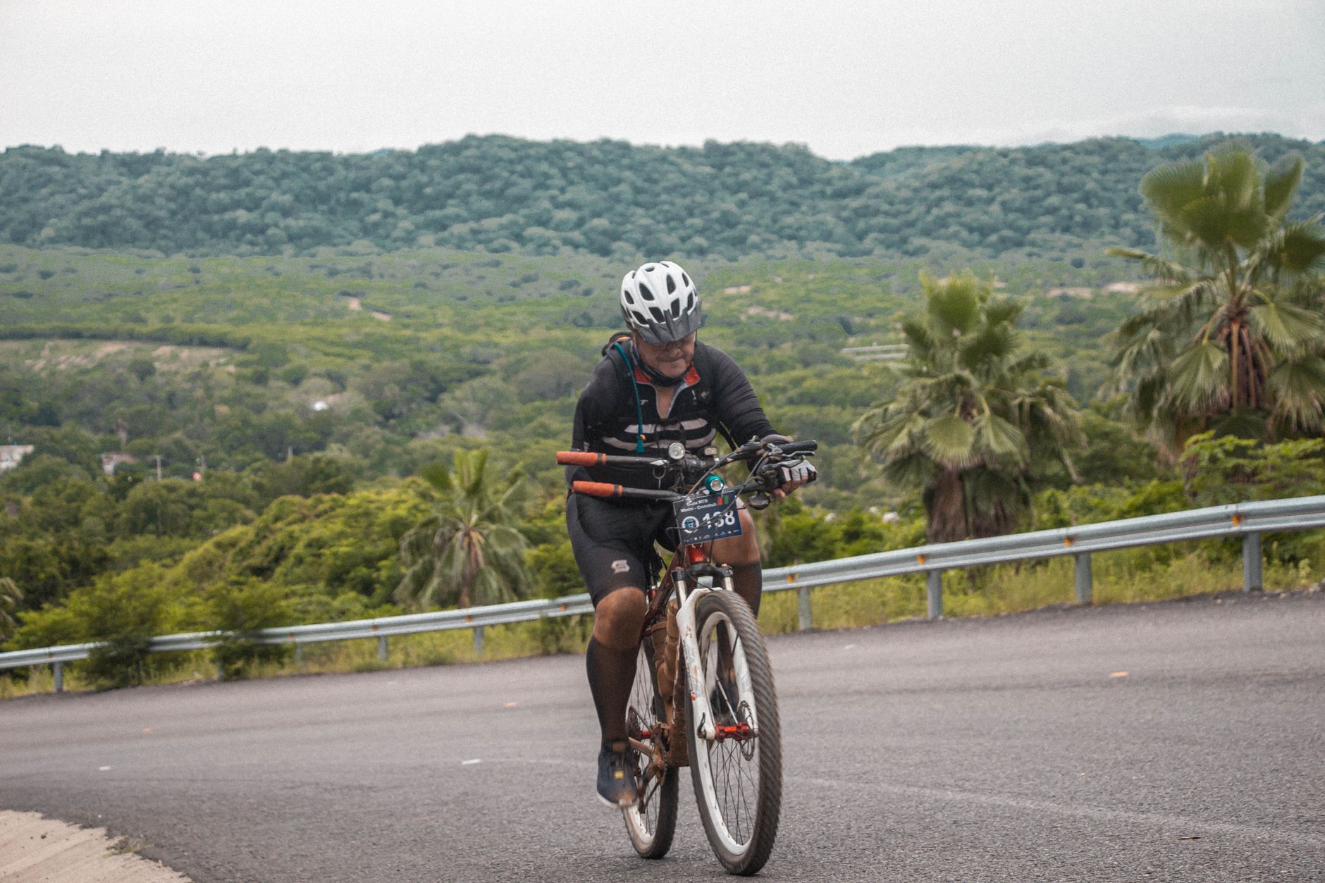 An adult male mountain biker riding uphill on a scenic road surrounded by lush greenery.