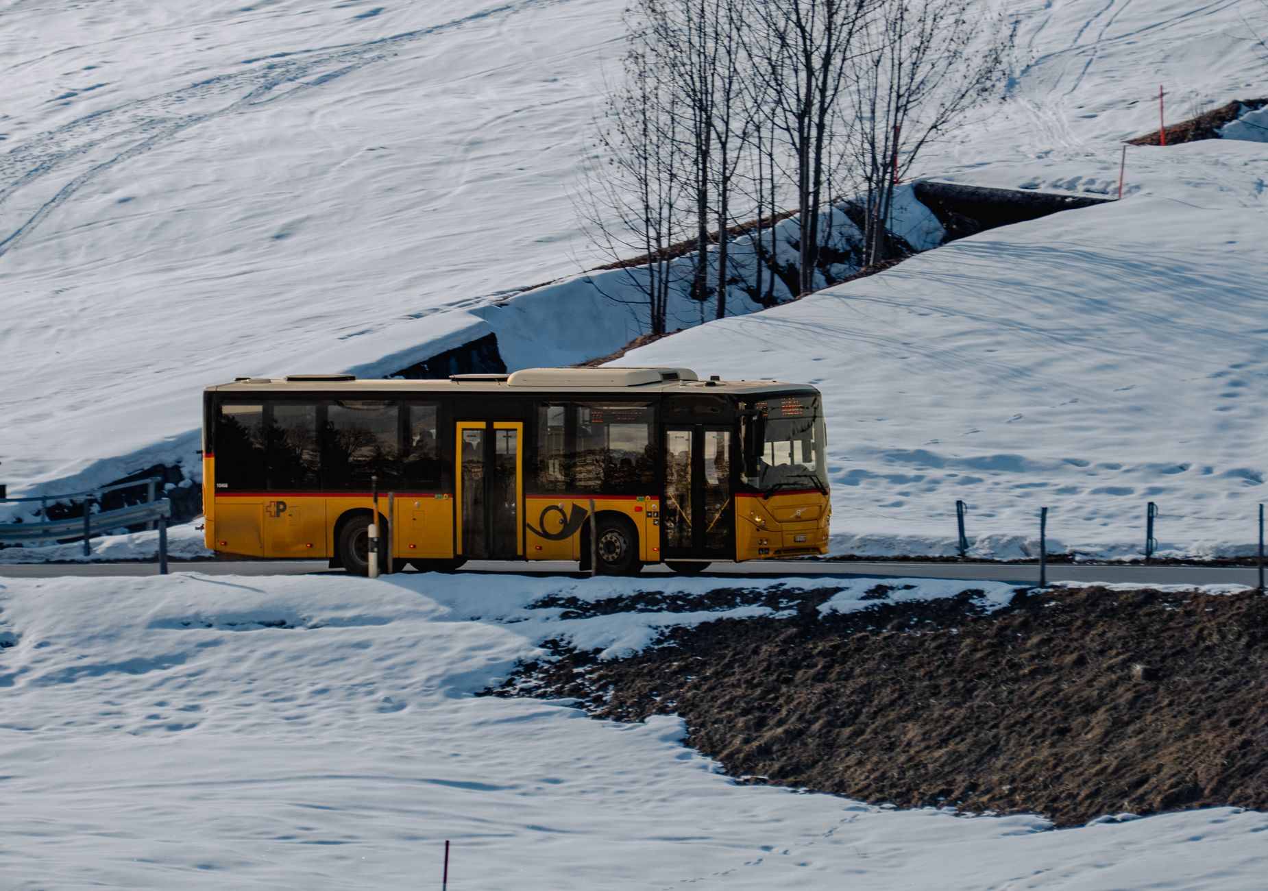 A yellow bus travels along a snowy mountain road, showcasing winter's serene beauty.