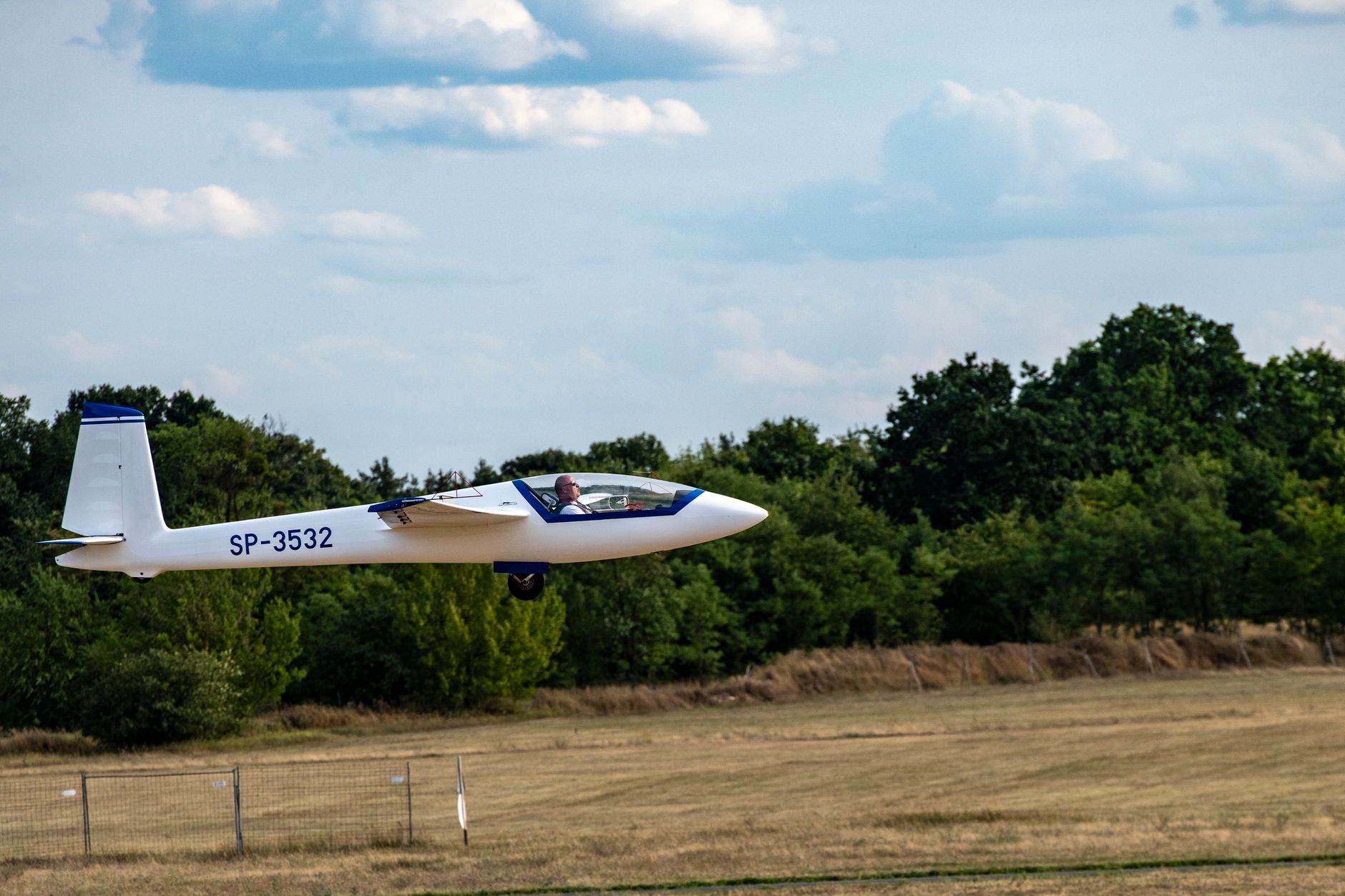 A glider plane in flight, soaring over a grassy airfield with trees in the background.