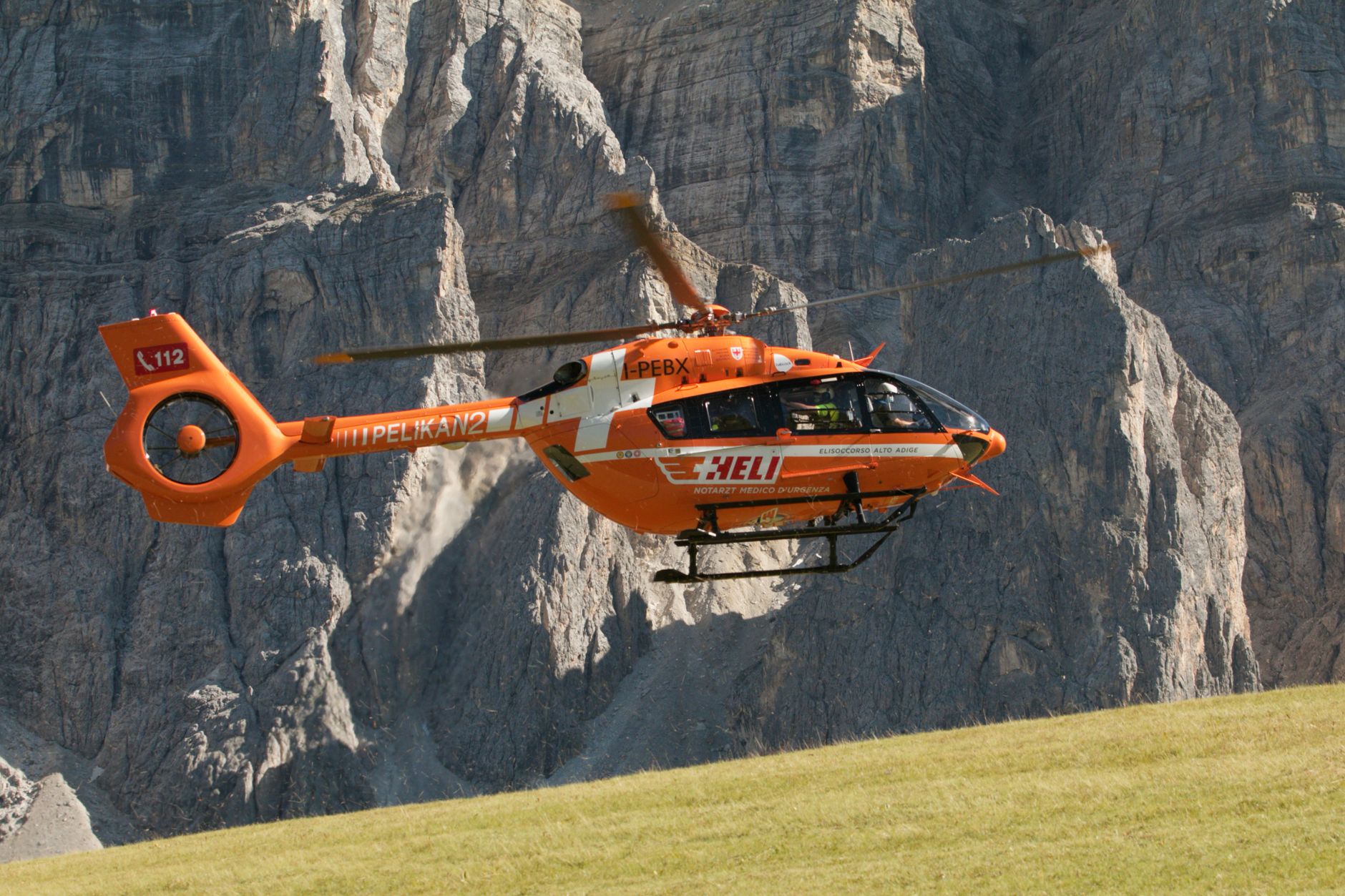Orange rescue helicopter in flight over Campill's mountainous terrain in Trentino-Alto Adige, Italy.