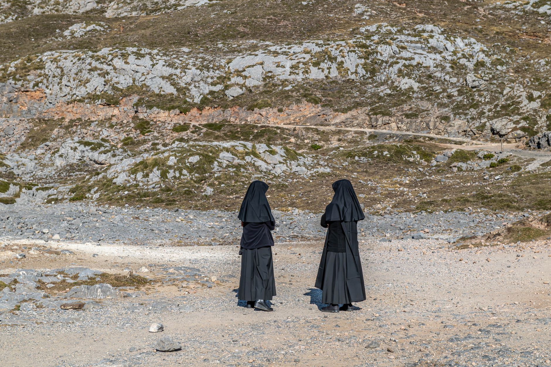 Two nuns in black habits walk in a rocky, remote landscape on a sunny day.