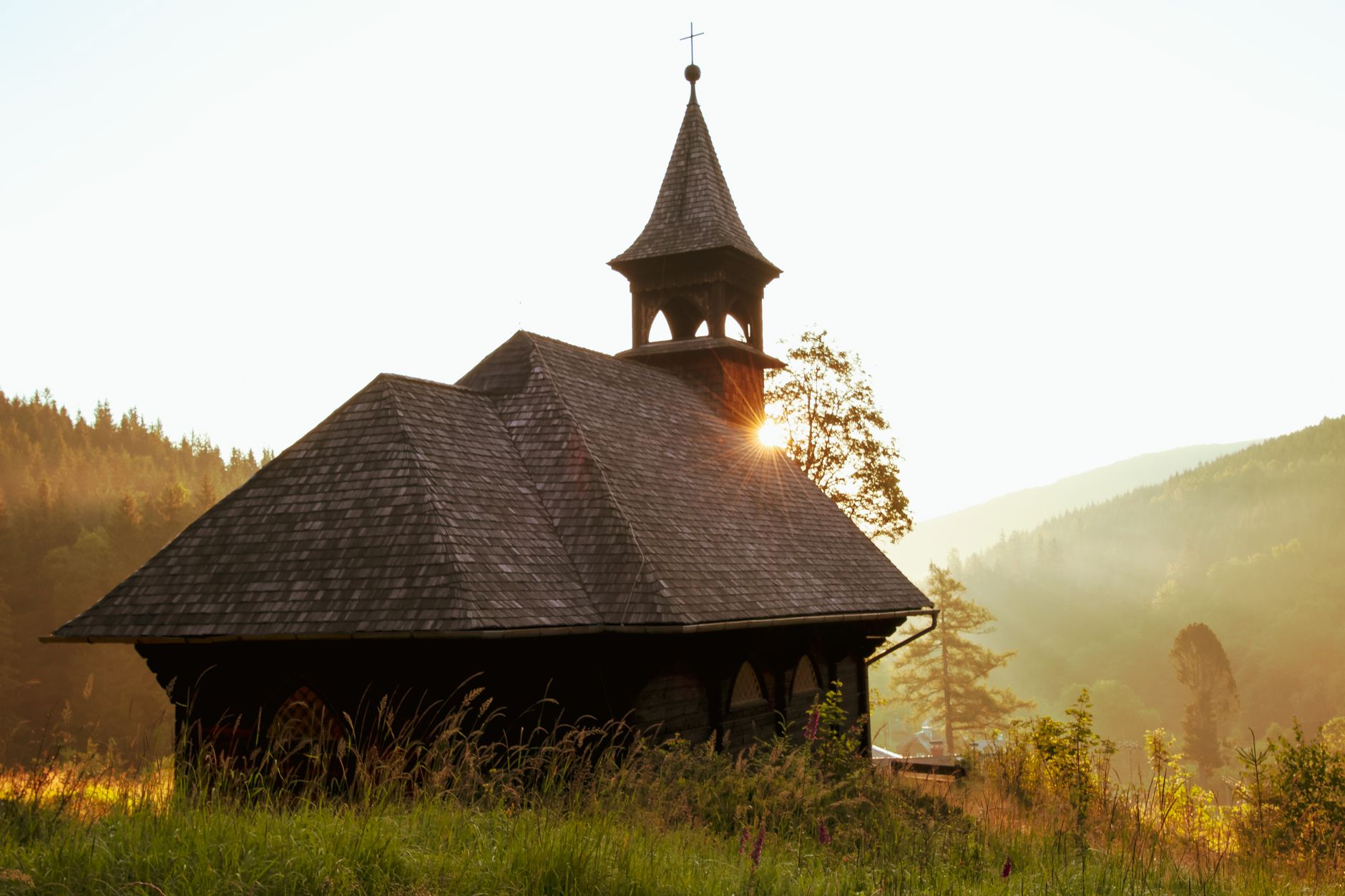 A serene rustic chapel with a tower glowing in sunset light amidst a lush mountain landscape.