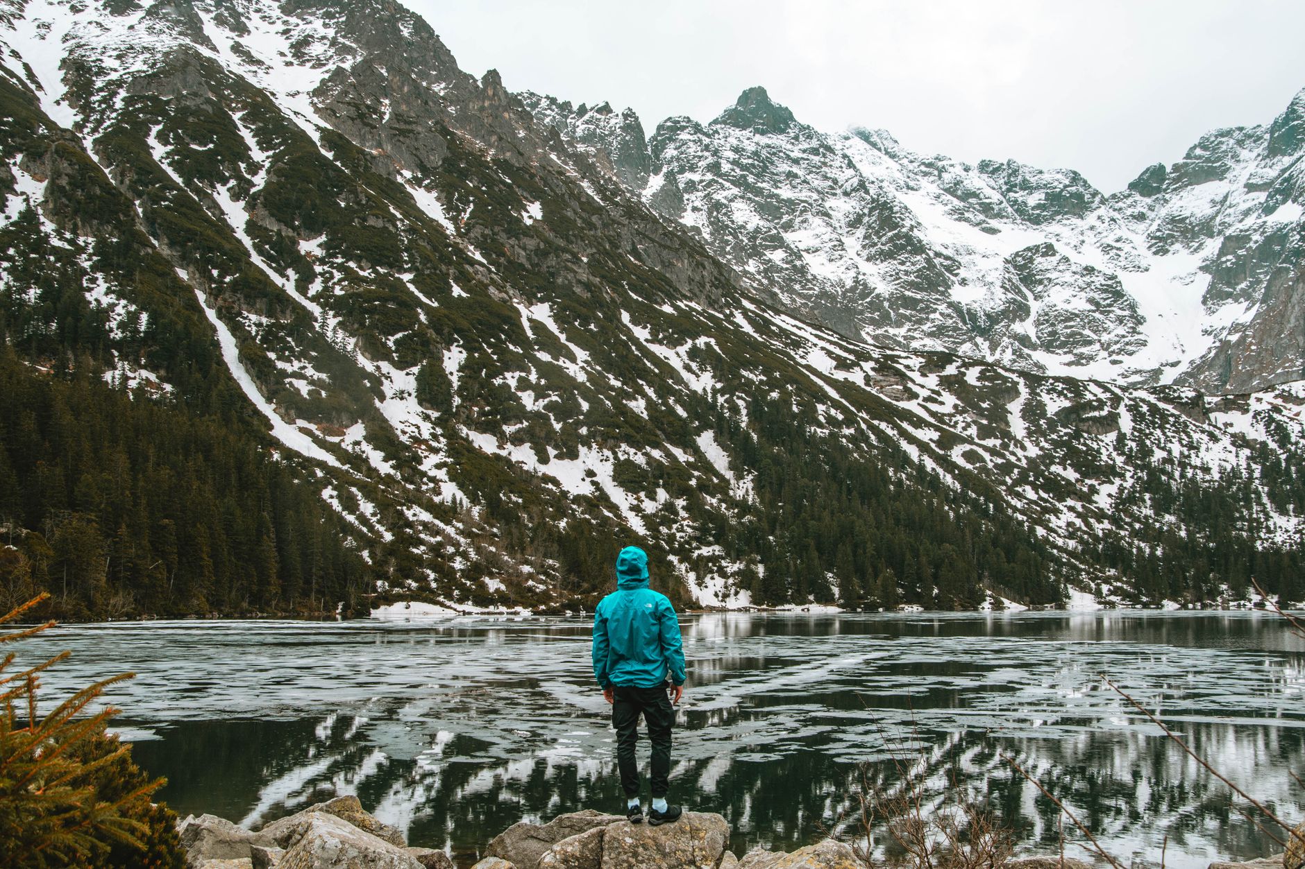 A solitary hiker gazes across a frozen mountain lake in winter, surrounded by snow-capped peaks.