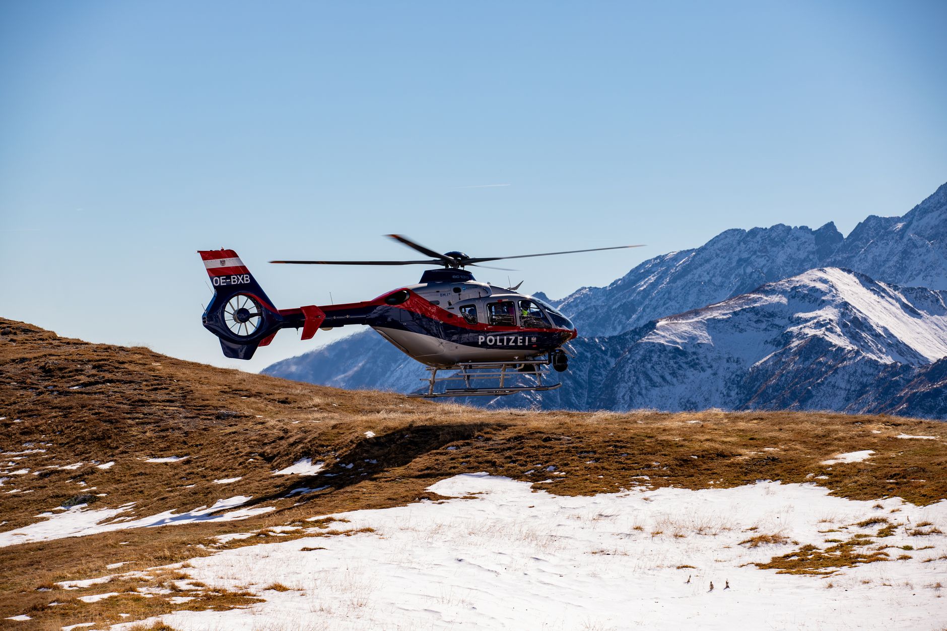 A police helicopter hovers above snowy alpine terrain, with rugged mountains in the backdrop.
