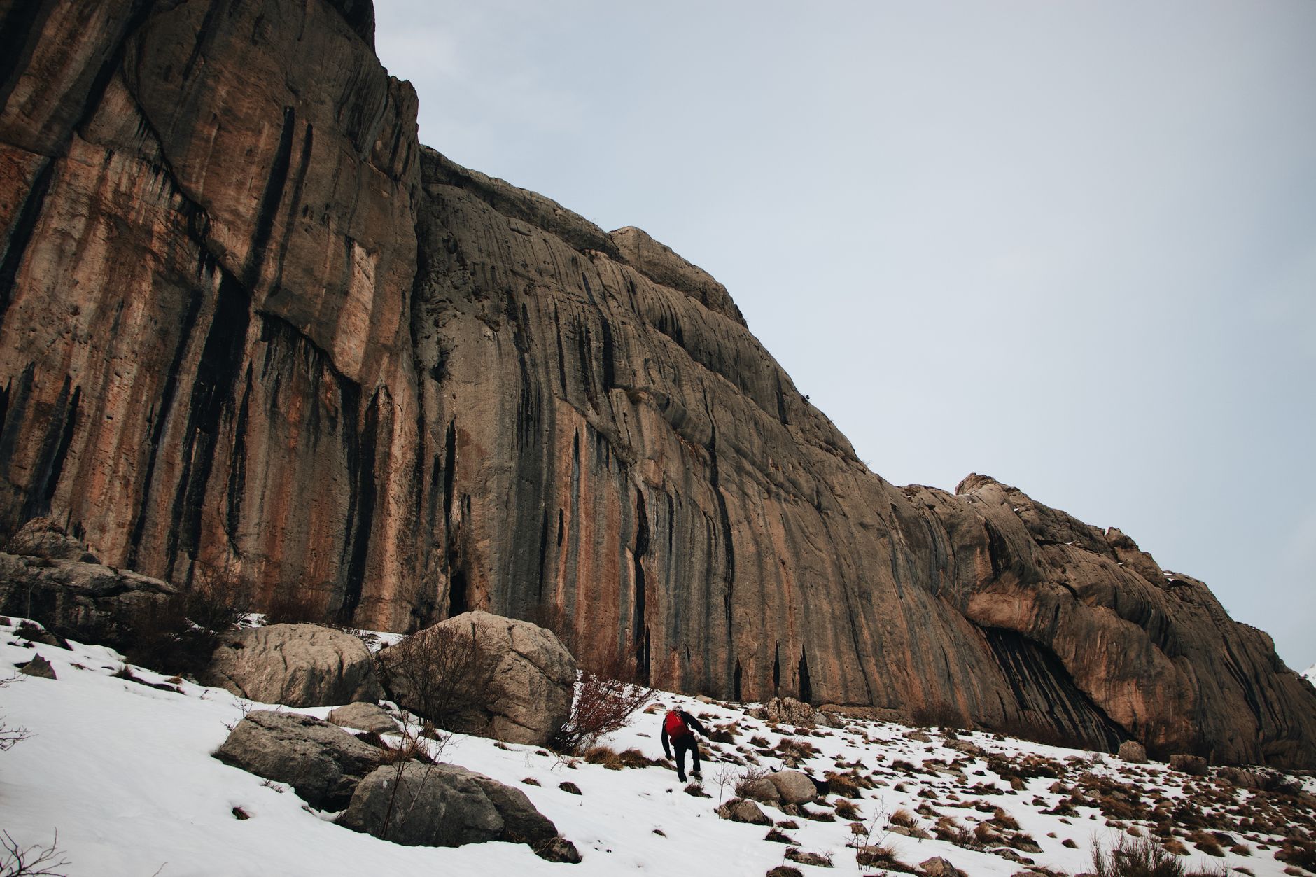 A lone hiker climbs a snow-covered mountain slope beneath rugged cliffs, set against a winter sky.