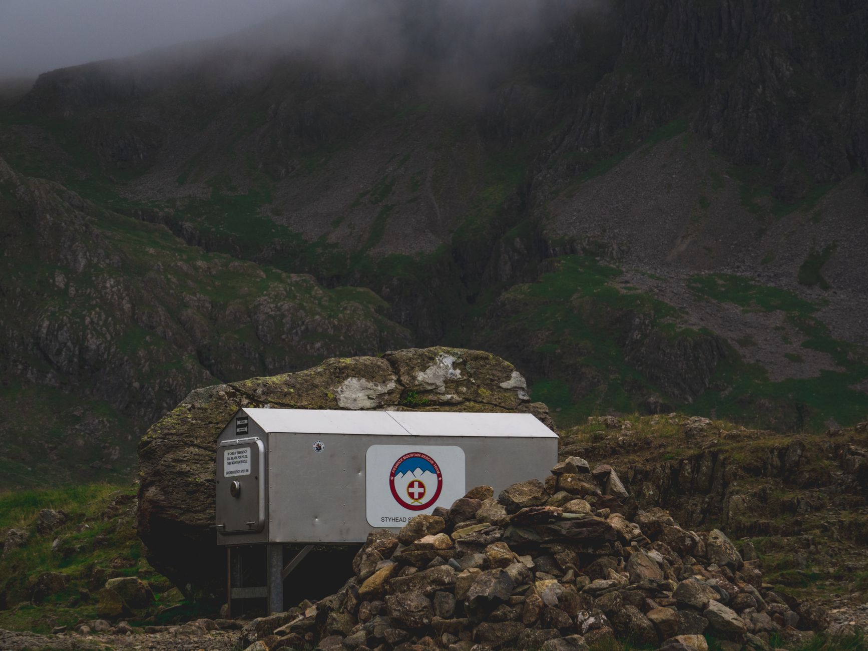 A mountain rescue shelter with stone cairns under dark, misty skies in rugged terrain.