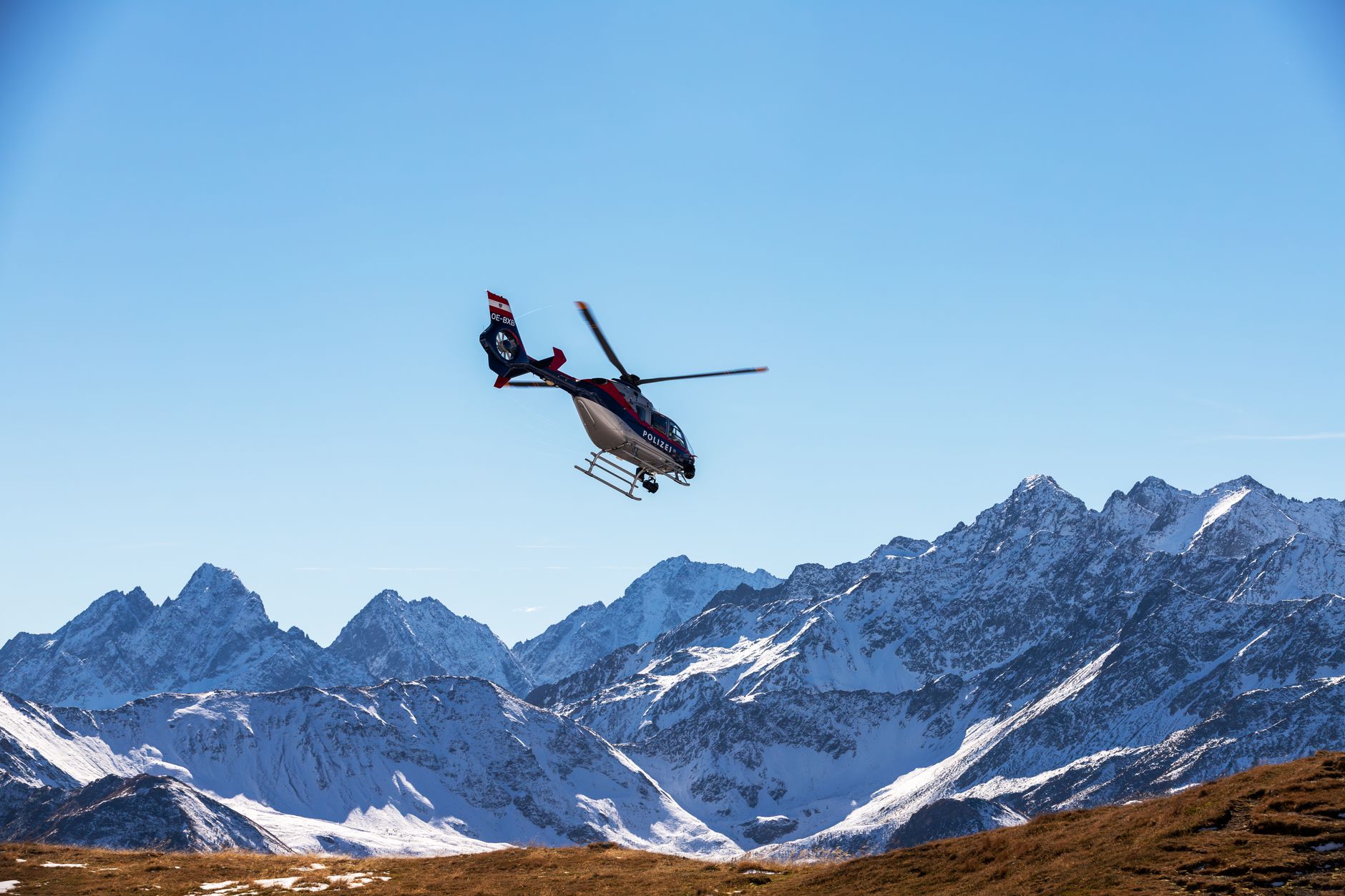 A police helicopter flies above snow-covered alpine mountains under a clear blue sky in Korutany, Austria.