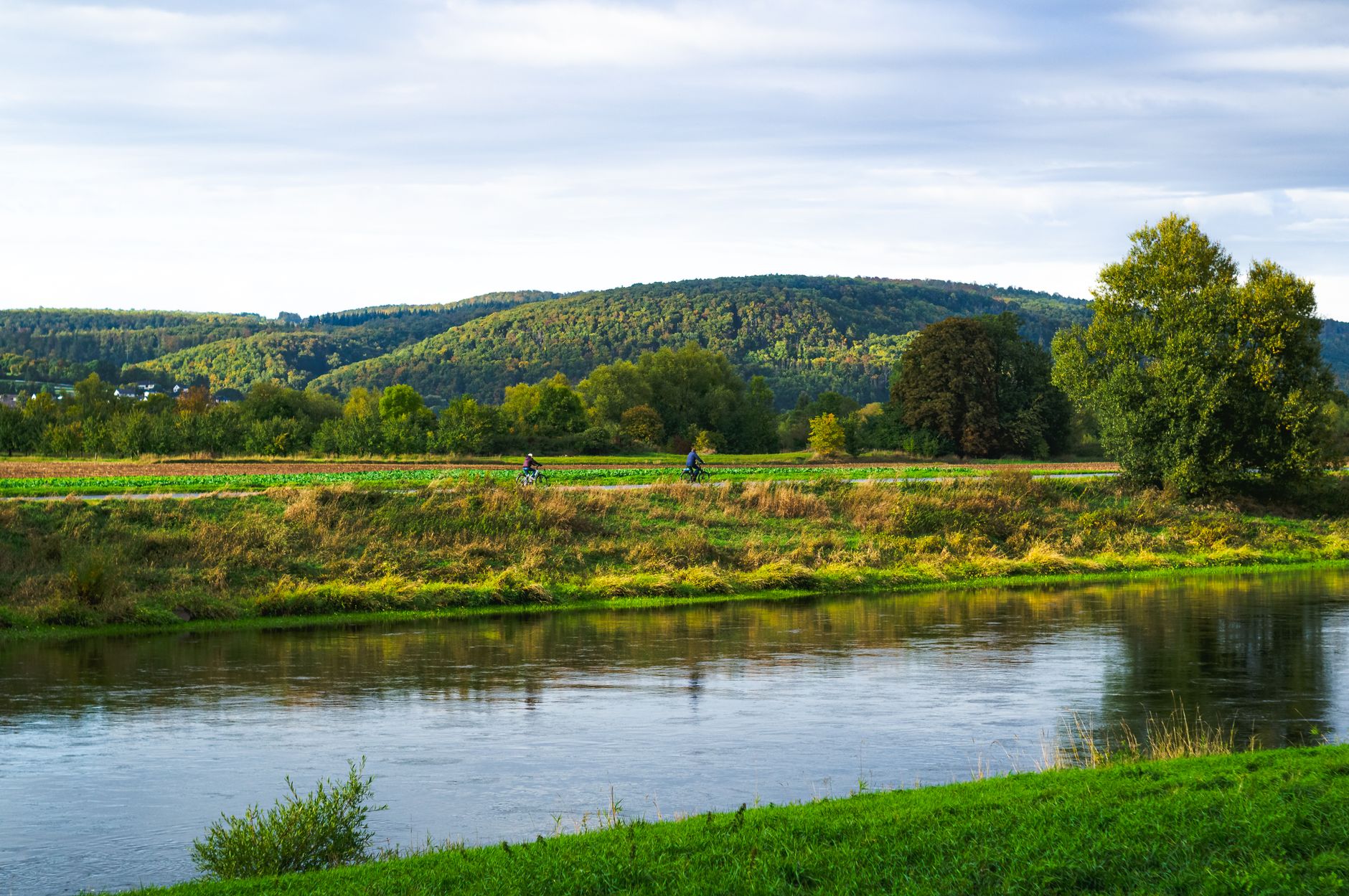 Two cyclists ride on a scenic riverbank path amidst lush green hills under a clear sky.